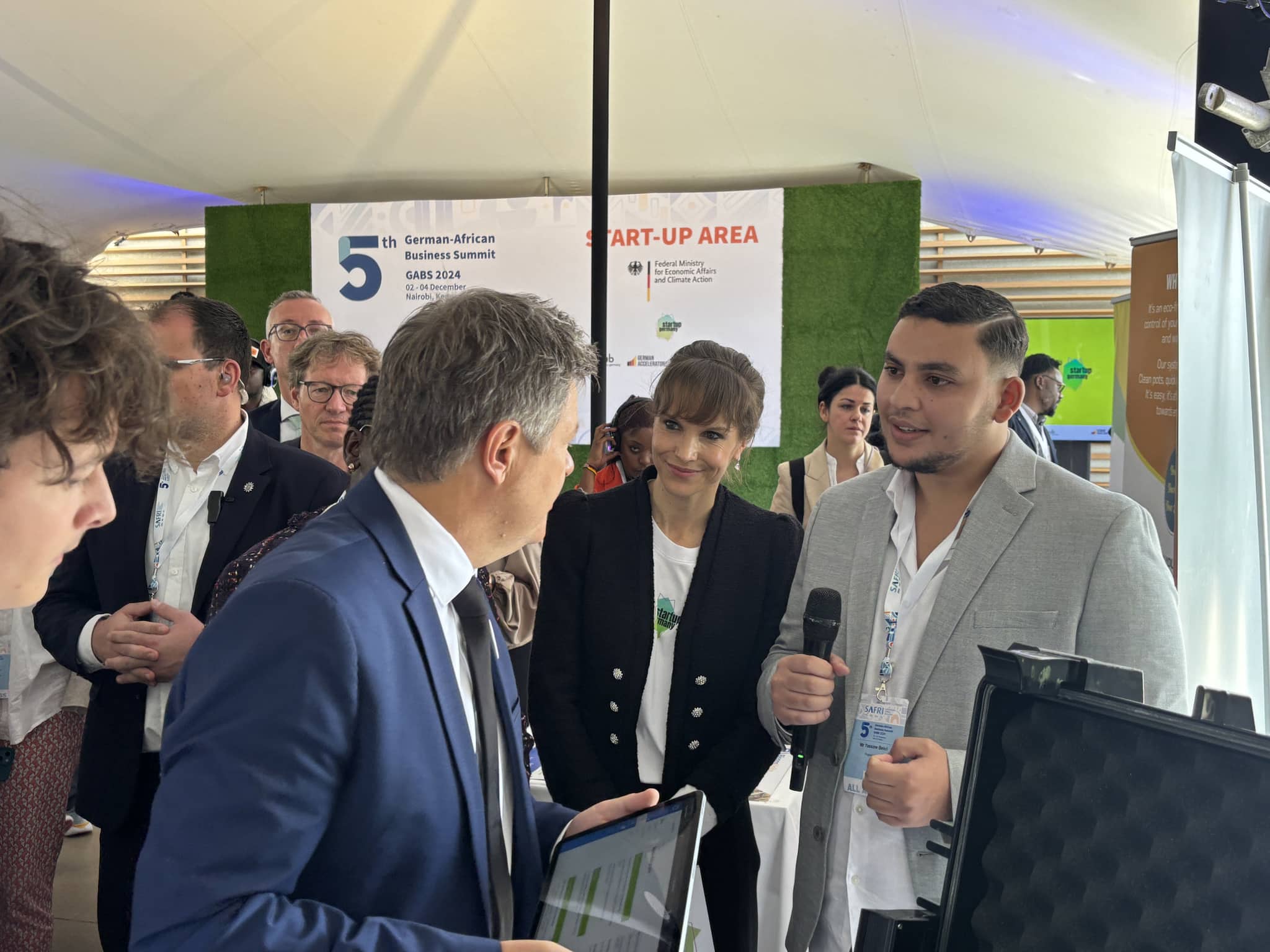 Yassine Bekri, Mikrofon in der Hand, zeigt dem damaligen Vizekanzler Robert Habeck das Tablet am Stand von Project Noor; dahinter das Banner des GABS 2024.