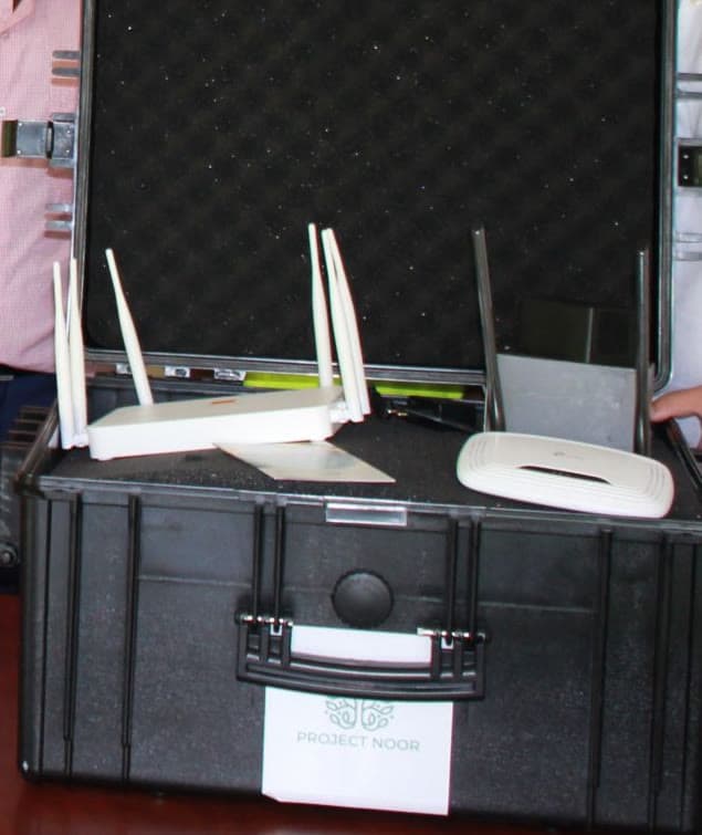 Ein geöffneter schwarzer Transportkoffer steht auf einem Tisch; im Schaumstoff liegen zwei WLAN-Router mit Antennen und ein Access Point, an der Front ein Schild mit dem Project-Noor-Zeichen. Am linken und rechten Bildrand stehen Personen.