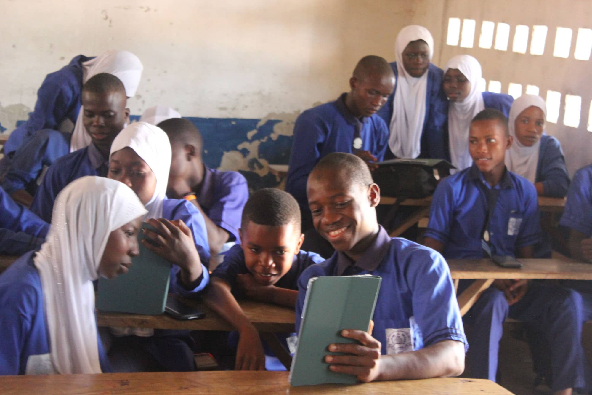 Ein Schüler lächelt auf das Tablet in seiner Hand, Mitschüler in blauer Uniform beugen sich zu ihm.